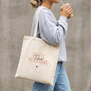 Könnte beinhalten: Beige Tragetasche mit den Worten "Beste Tina Hebamme" in rosa und schwarzer Schrift, mit einem kleinen schwarzen Herz. Die Tasche hat lange Träger und wird über der Schulter getragen.