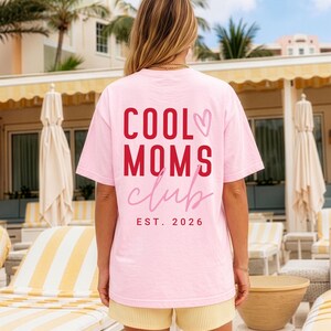 Könnte beinhalten: Ein hellrosa T-Shirt mit dem roten Schriftzug COOL MOMS, dem rosafarbenen kursiven Schriftzug club, einem rosa Herzumriss und dem roten Schriftzug EST. 2026 auf der Rückseite. Das T-Shirt hat kurze Ärmel und einen Rundhalsausschnitt.
