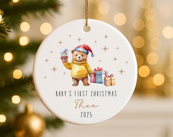 May include: A white ceramic Christmas ornament with a watercolor illustration of a teddy bear wearing a Santa hat and holding a snow globe. The text reads "Baby's First Christmas" and the name "Theo" with the year "2025".