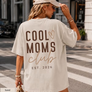 Könnte beinhalten: Ein sandfarbenes Kurzarm-T-Shirt mit dem dunkelbraunen Schriftzug COOL MOMS club, einem Herzumriss und dem Text EST. 2026 auf der Rückseite. Das T-Shirt wird mit einer passenden sandfarbenen Baseballkappe und einer Handtasche kombiniert.