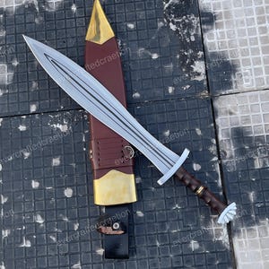 May include: A silver sword with a brown handle and a leather sheath. The sword has a double-edged blade and a crossguard. The sheath is brown with gold accents. The sword is resting on a dark, textured surface.