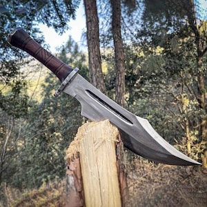 May include: A large, silver-colored knife with a dark brown wooden handle is embedded in a wooden post. The knife has a wide blade with a curved edge and a decorative handle. The background features trees and foliage.
