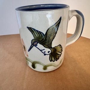 Puede incluir: Taza de cerámica color crema con borde y asa azules. La taza presenta un colibrí pintado a mano en vuelo, con detalles verdes y azules. Una flor rosa y enredaderas verdes también están pintadas en la taza.