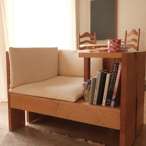 May include: A wooden bench with an integrated side table. The bench features a light-coloured cushion and a shelf with books and pens. A red and white patterned mug sits on the table. The room has a neutral colour palette.