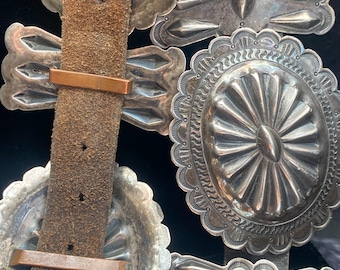 May include: A silver belt buckle with intricate designs. The buckle is made up of several pieces, each with a different pattern. The buckle is attached to a brown leather belt.