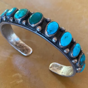 May include: A silver cuff bracelet featuring a row of turquoise stones. The stones are set in silver and surrounded by small silver beads.