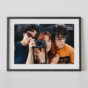 May include: A framed photograph featuring three people, one holding a black camera. The person in the center is looking through the camera lens. The person on the right is wearing an orange shirt with the text "ECT OF DESIRE".