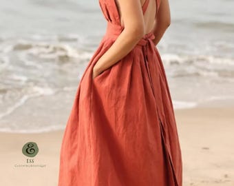 May include: A rust-colored linen dress with a cross-back design and a flowing skirt. The dress features pockets and a tie-back detail. The image is taken on a beach, with the ocean in the background. The dress is from ESS Clothing Boutique.