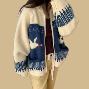 May include: Cream-colored zip-up cardigan with navy blue geometric and bird pattern details. The sweater has two front pockets and a cozy, oversized fit. The sleeves and lower hem feature a navy blue pattern. A casual, comfortable outerwear option.