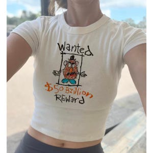 Può includere: Maglietta bianca corta con un disegno animato di Mr. Potato Head in un manifesto di ricerca. Il testo dice "Wanted" e "$50 bzillion Reward".