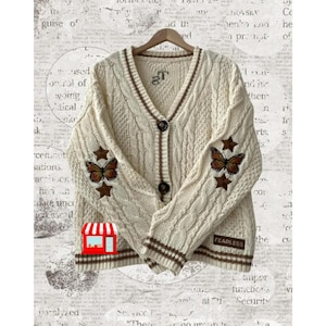 May include: A cream-coloured cable knit cardigan with brown trim and embroidered butterflies on the sleeves. The cardigan has a button closure and the text "FEARLESS" is embroidered on the bottom right side.