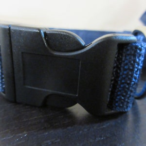 May include: Close-up of a black plastic dog collar buckle with a blue fabric strap. The buckle is a quick-release type, with a curved design. A silver metal ring is visible on the side.