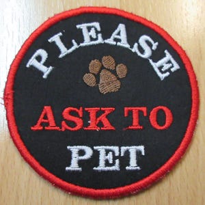 May include: Embroidered circular patch with a black background and a red border. The patch features the words "PLEASE ASK TO PET" in white and red lettering, with a brown paw print in the center. This patch is designed for pet lovers.