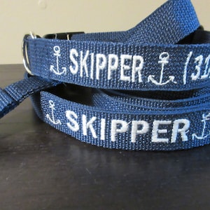 May include: Navy blue dog collar and leash set. The collar and leash are embroidered with the name "SKIPPER" in white, along with anchor designs. The leash has a silver-colored metal clasp.