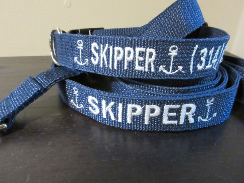 May include: Navy blue dog collar and leash set. The collar and leash are embroidered with the name "SKIPPER" in white, along with anchor designs. The leash has a silver-colored metal clasp.