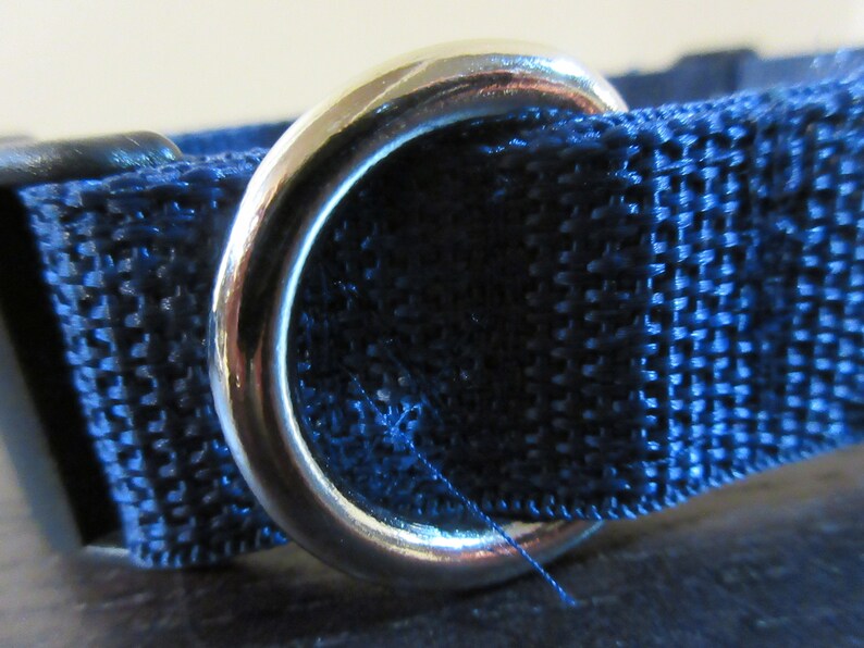 May include: Close-up of a navy blue nylon webbing with a shiny silver metal ring. The webbing has a textured weave and is stitched together. The ring is smooth and reflective, contrasting with the fabric.