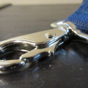 May include: Close-up of a shiny, silver-colored metal clasp with a curved hook and a spring-loaded lever. A blue woven strap is attached to the clasp. The clasp is likely part of a leash or strap.