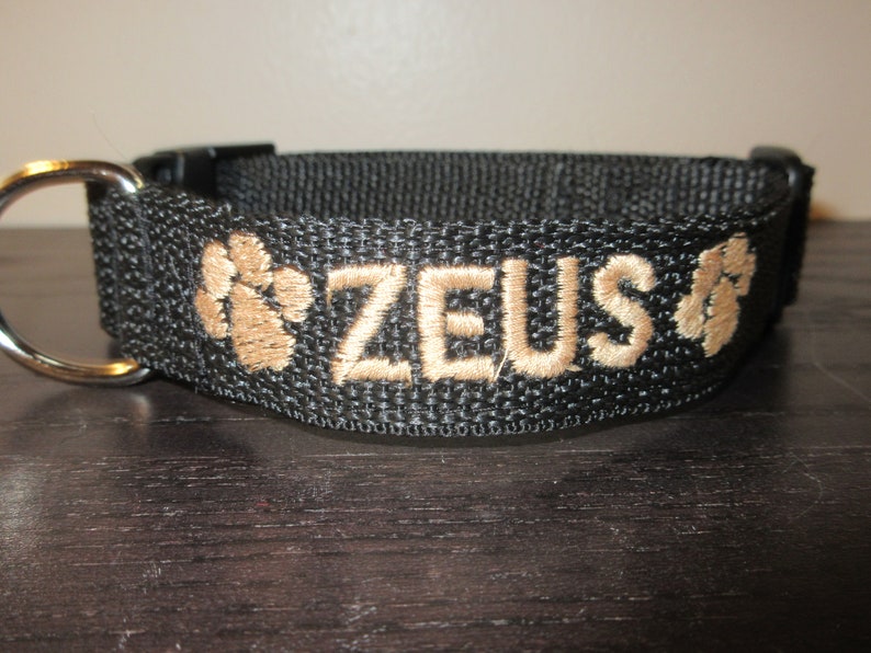 May include: Black dog collar with the name "ZEUS" embroidered in tan thread. The collar features tan paw print designs on either side of the name. Includes a metal D-ring and a black plastic buckle.