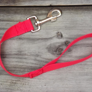May include: A bright red nylon dog leash featuring a silver-toned metal swivel snap hook at one end and a looped handle at the other. The durable woven fabric strap is stitched securely at the connection points for added strength and stability.