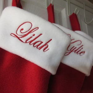 Peut inclure: Une série de chaussettes de Noël rouges avec des revers en peluche blanche. Chaque chaussette présente un prénom brodé en écriture cursive rouge sur le revers blanc, incluant Lilah et Kylie, avec des boucles en tissu rouge pour suspendre.