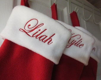 May include: A series of red Christmas stockings with white plush cuffs. Each stocking features a name embroidered in red cursive script on the white cuff, including the names Lilah and Kylie, with red fabric loops for hanging on white hooks.