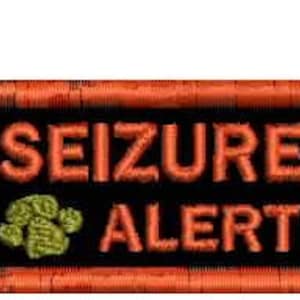 Peut inclure: Écusson brodé avec les mots "SEIZURE ALERT" en fil orange sur fond noir. Un motif de patte verte est à gauche. L'écusson a une bordure orange.