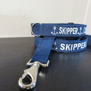 May include: Navy blue dog collar and leash set. The leash and collar are made of a woven material with the word "SKIPPER" and an anchor embroidered in white. The leash has a silver-colored metal clasp.
