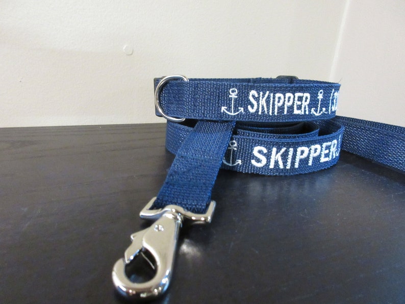 May include: Navy blue dog collar and leash set. The leash and collar are made of a woven material with the word "SKIPPER" and an anchor embroidered in white. The leash has a silver-colored metal clasp.