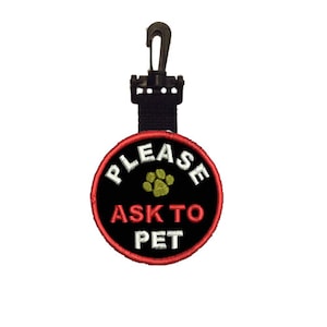 Puede incluir: Una etiqueta circular negra y roja con el texto "PLEASE ASK TO PET" en blanco y una huella de pata verde. La etiqueta está sujeta a una correa negra con un clip negro.