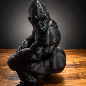May include: A black gorilla figurine in a crouching position, showcasing detailed muscular anatomy and textured fur patterns across its body. The sculpture is rendered in a solid matte black finish, capturing the primate's powerful build and focused facial expression while it sits in a low, balanced stance.