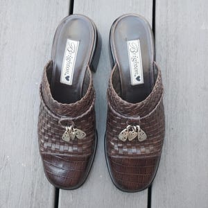 May include: A pair of brown leather clogs with a woven design on the upper. The clogs have a small heel and decorative silver charms. The brand name "Brighton" is visible on the insole.