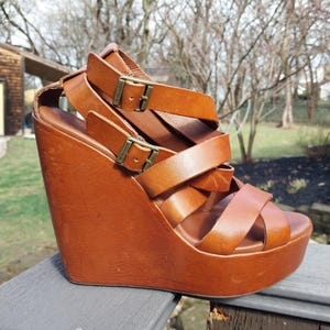 May include: Brown leather wedge sandals with multiple straps and buckle closures. The platform sole and wedge heel are also brown. The sandals are sitting on a gray wooden surface.