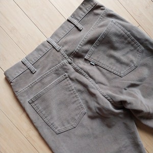 May include: Close-up of the back of a pair of taupe corduroy pants. The pants have two back pockets, belt loops, and a small tag. The fabric has a textured appearance.