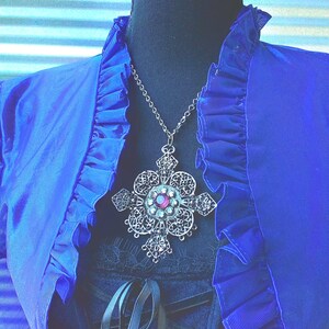 May include: A silver-toned filigree pendant necklace with a large cross design, featuring a central pink gemstone. The necklace hangs against a blue ruffled top, creating a striking contrast. The pendant's intricate details and the top's texture are highlighted.