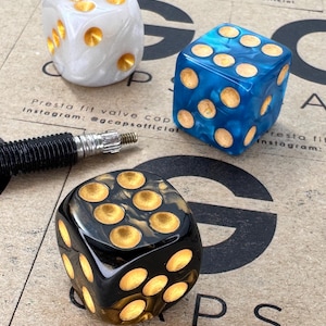 Puede incluir: Tres dados en blanco, azul y negro con puntos dorados. Los dados están sobre una superficie marrón con el texto "CAPS" y "Presta fit valve caps Instagram: @gcapsofficial".
