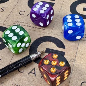 May include: A collection of colourful dice in green, purple, blue, and red with white and gold pips. A black valve stem is also present. The dice are displayed on a brown surface with the text "Presta fit valve caps" and an Instagram handle.