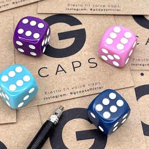 May include: Four dice valve caps in purple, pink, blue, and navy blue, each with white dots. The caps are arranged on a brown surface with the text "G CAPS" and "Presta fit valve caps" visible.