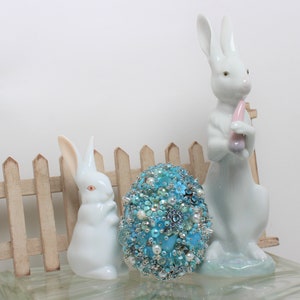 May include: Two white porcelain bunny figurines stand beside a blue and white beaded Easter egg. The bunnies are holding a pink and white carrot and a blue and white egg. The egg is decorated with beads, sequins, and pearls.