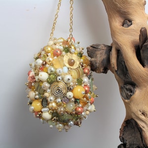 May include: A gold chain hangs from a large, round, beaded ornament. The ornament is made of a variety of materials, including glass beads, metal beads, and shells. The ornament is a light beige, gold, and green color.