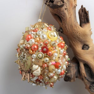 May include: A gold and white beaded ornament with a variety of colors, including peach, coral, and pink. The ornament is shaped like an egg and is hanging from a branch.