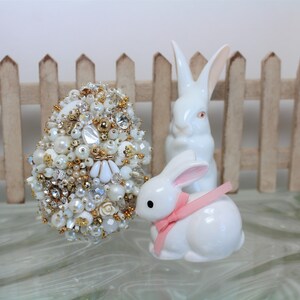 May include: A white ceramic Easter egg decorated with pearls, rhinestones, and gold beads. Two white ceramic bunnies are beside the egg, one with a pink ribbon around its neck.