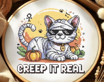 Puede incluir: Bordado de punto de cruz en un aro de madera. El diseño presenta un gato disfrazado de momia, con gafas de sol y un collar dorado con un dije de calavera. Incluye una calabaza y el texto "CREEP IT REAL", sobre un fondo amarillo.