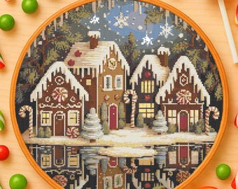 May include: A cross-stitch embroidery of a winter village scene, featuring gingerbread houses with snow-covered roofs and candy cane accents. The artwork is set in a wooden embroidery hoop, surrounded by colorful candies and a lollipop.