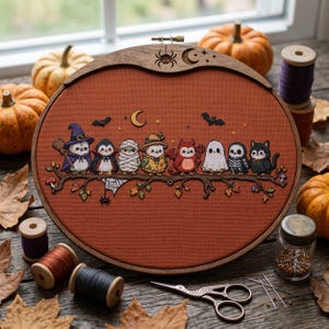 May include: A round wooden embroidery hoop containing a cross-stitch design on rust-colored fabric. The pattern features eight small creatures dressed in Halloween costumes, including a witch, vampire, mummy, and ghost, perched on a tree branch.