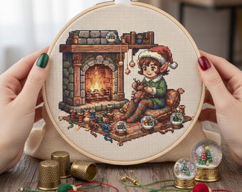 Könnte beinhalten: Ein Kreuzstich-Stickrahmen mit einer Weihnachtsszene mit einem Elf am Kamin. Der Elf trägt ein grünes Outfit und einen Weihnachtsmütze. Die Szene umfasst Schneekugeln, Garnrollen und einen Teppich. Der Rahmen wird von Händen mit grünem und rotem Nagellack gehalten.