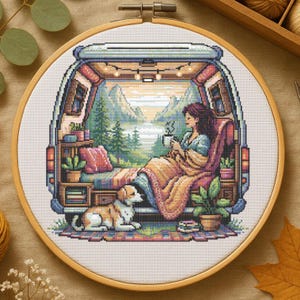 May include: Cross-stitch embroidery in a wooden hoop depicts a cozy camper interior with a woman sipping coffee, a dog, and a scenic mountain view. The scene is detailed with plants, a blanket, and string lights, creating a warm, inviting atmosphere.