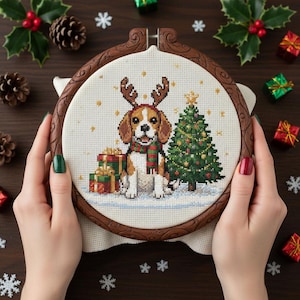 May include: A cross-stitch embroidery hoop featuring a beagle wearing reindeer antlers and a scarf, with a Christmas tree and presents. The hoop is held by hands with green and red nail polish. The background includes pine cones, holly, and small wrapped gifts.