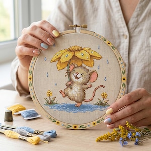 以下が含まれることがあります： 黄色い花の傘の下で雨の中を歩く、陽気なネズミが描かれたクロスステッチ刺繍枠。木製の枠は花柄で装飾されています。刺繍糸と指ぬきがテーブルに置かれています。