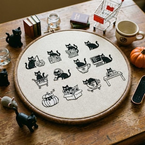 May include: A round, wooden embroidery hoop with a cream-colored fabric featuring black cross-stitch cat designs. The cats are depicted in various scenarios, such as reading, using a computer, and riding a skateboard. The hoop is surrounded by miniature objects.