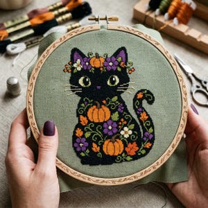 May include: A cross-stitch embroidery project in a round wooden hoop featuring a black cat with large green eyes. The cat is decorated with orange pumpkins, purple flowers, and green leaves on a sage green fabric background.